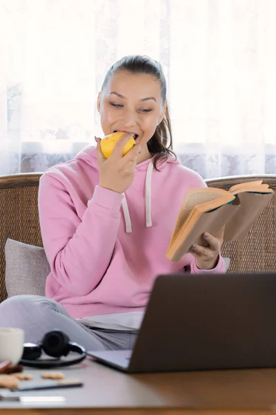 Chica sonriente trabaja de forma remota con ordenador portátil comer limón y leer libro — Foto de Stock