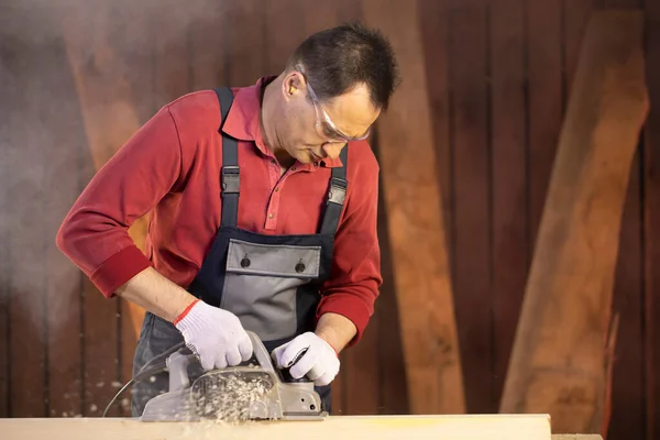 Middle-aged male craftsman in googles treats piece of wood with electric planer — Stock Photo, Image