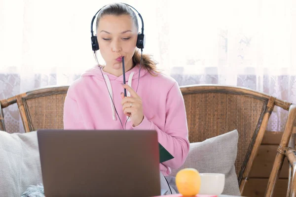 Chica en sudadera con capucha rosa y auriculares cuidadosamente mirando portátil lápiz de mascar — Foto de Stock