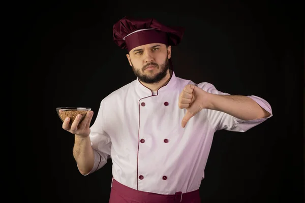 Hombre chef insatisfecho en uniforme sostiene tazón con trigo sarraceno muestra signo de aversión —  Fotos de Stock
