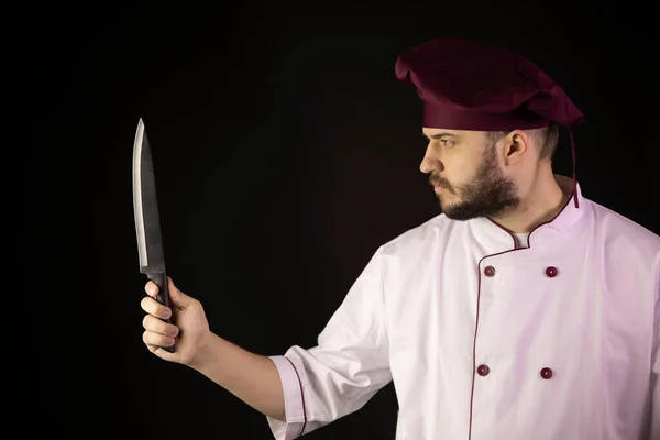Joven guapo barbudo chef hombre en uniforme sostiene cuchillo en la mano mira fijamente —  Fotos de Stock