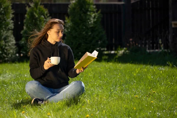 Siyah kapüşonlu çekici esmer çimen üzerinde oturmuş kitap okuyor, bir fincan kahve içiyor. — Stok fotoğraf