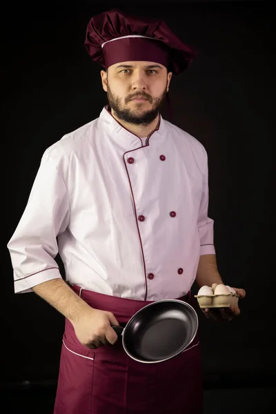 Jonge knappe bebaarde chef-kok in uniform houdt koekenpan en eieren — Stockfoto