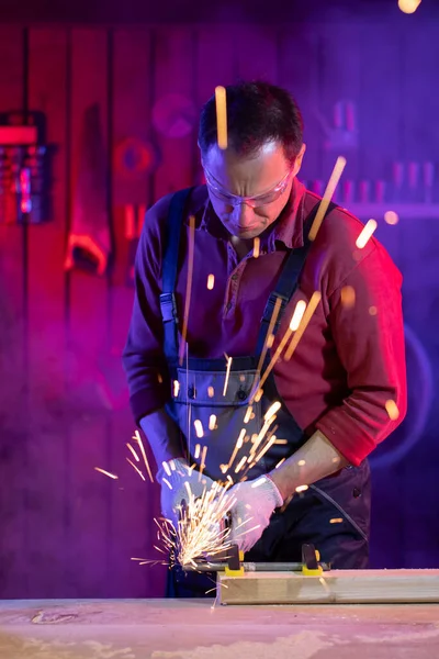 Man in jumpsuit goggles and gloves cutting metal with sparks in coloured light — Stock Photo, Image