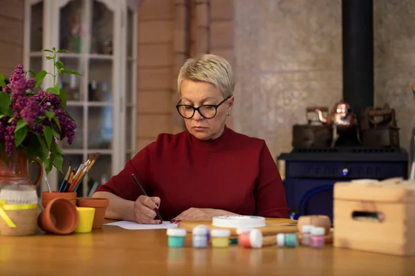 Μεσήλικη γυναίκα ζωγραφίζει με μολύβι σε άνετο εξοχικό σπίτι με τζάκι — Φωτογραφία Αρχείου