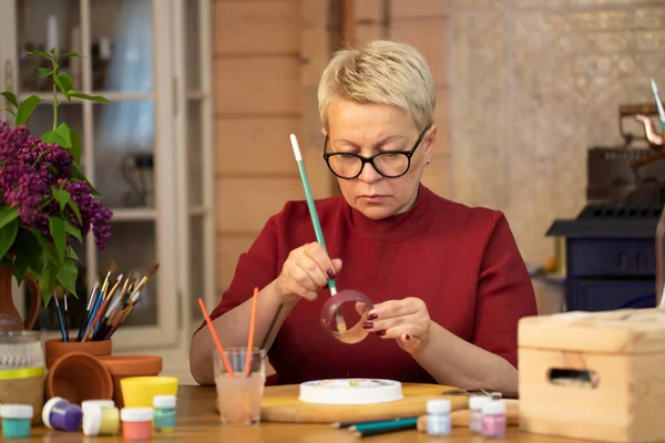 Mulher de meia-idade aguda pinta pulseira de madeira na casa de campo. passatempo predileto de desenho . — Fotografia de Stock
