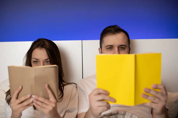 Beautiful couple lays in bed with paper books in hands hiding behind open books