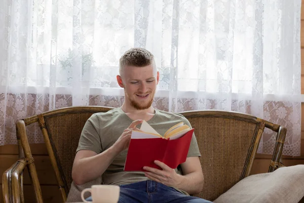 Kırsaldaki ahşap evde hasır bankta kitap okuyan gülümseyen genç adam. — Stok fotoğraf