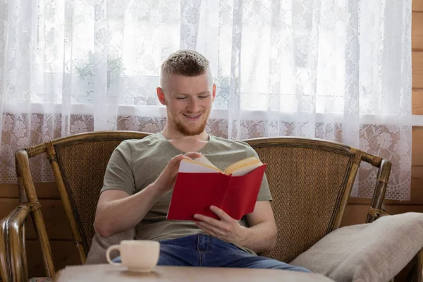 Genç adam ahşap evde kırmızı kapaklı kitap okuyor. — Stok fotoğraf