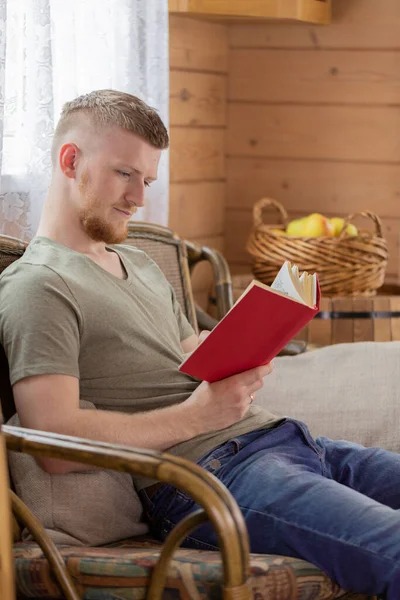 田舎の家のウィッカー・ベンチに赤い表紙の本を読んでいる若い男 — ストック写真