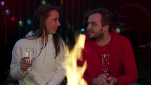Νεαρό ευτυχισμένο ζευγάρι που αναπαύεται κοντά σε φωτιά και πίνει σαμπάνια μια καλοκαιρινή νύχτα — Αρχείο Βίντεο