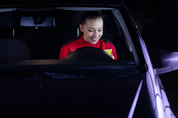 Sonriente morena pelo largo chica en coche conducción por la noche sonriendo leer mensajes — Foto de Stock