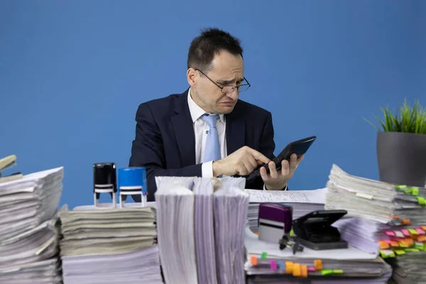 worried businessman counts on calculator at office desk with piles of papers