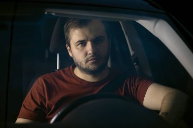 Erkek sürücü gece direksiyonda oturur düşünceli bir şekilde kameraya bakar.