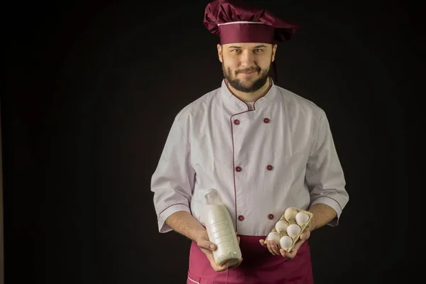 Chef sonriente hombre en uniforme sostiene botella de leche y huevos fondo negro —  Fotos de Stock