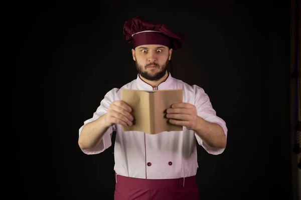 Verrast mannelijke kok in schort op recept-boek over zwarte achtergrond — Stockfoto