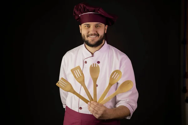 Positieve knappe bebaarde chef-kok in uniform houdt houten keukengerei — Stockfoto