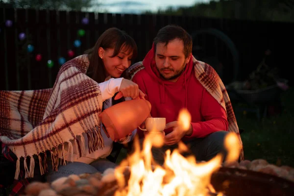 couple of blanketed lovers by bonfire pouring coffee from thermos into cup