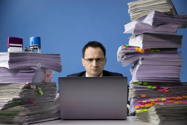 Homem de negócios trabalha em laptop de computador na mesa de escritório com enorme pilha de documentos — Fotografia de Stock