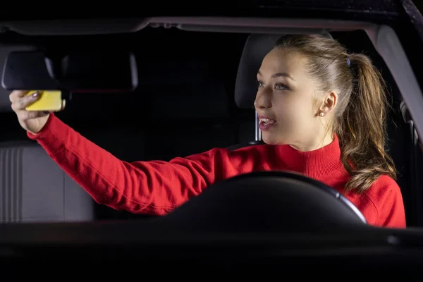 Linda chica morena de pelo largo en suéter rojo coche de conducción por la noche hace selfies — Foto de Stock