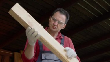 Koruyucu gözlüklü marangoz elinde tahta bir kiriş tutuyor ve onu inceliyor.