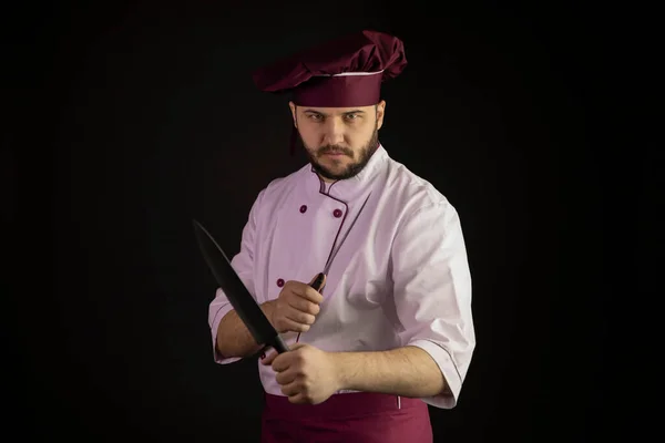 Handsome bearded chef cook in violet apron and cap holds crossed sharp knives — Stock Photo, Image