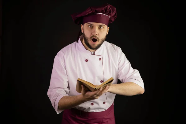 Verbaasd bebaarde chef-kok in uniform houdt open boek van vintage oude recepten — Stockfoto