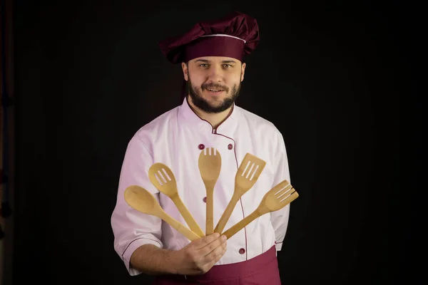 smiling young bearded male chef cook in uniform holding wooden kitchen utensils