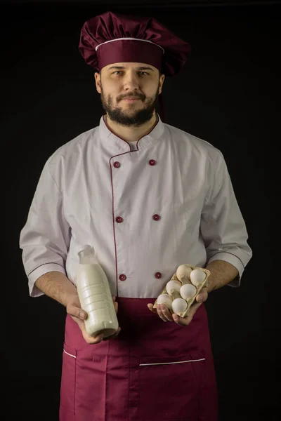 Бородатий майстер шеф-кухар в уніформі тримає пляшку молока і півдюжини яєць — стокове фото