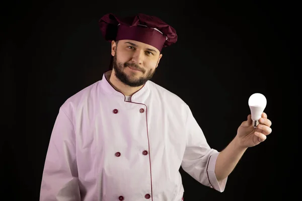 Бородатий майстер шеф-кухар в уніформі тримає енергозберігаючу лампочку, дивлячись на камеру — стокове фото