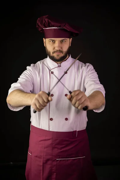 bearded male chef cook wearing violet apron and cap holds crossed sharp knives