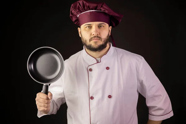 Майстер шеф-кухар в уніформі руків'я сковорідка дивиться на камеру ізольовані на чорному — стокове фото