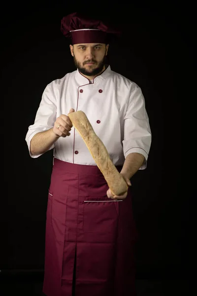 Panadero hombre vistiendo delantal violeta y la tapa mantiene pan Baguette aislado en negro —  Fotos de Stock