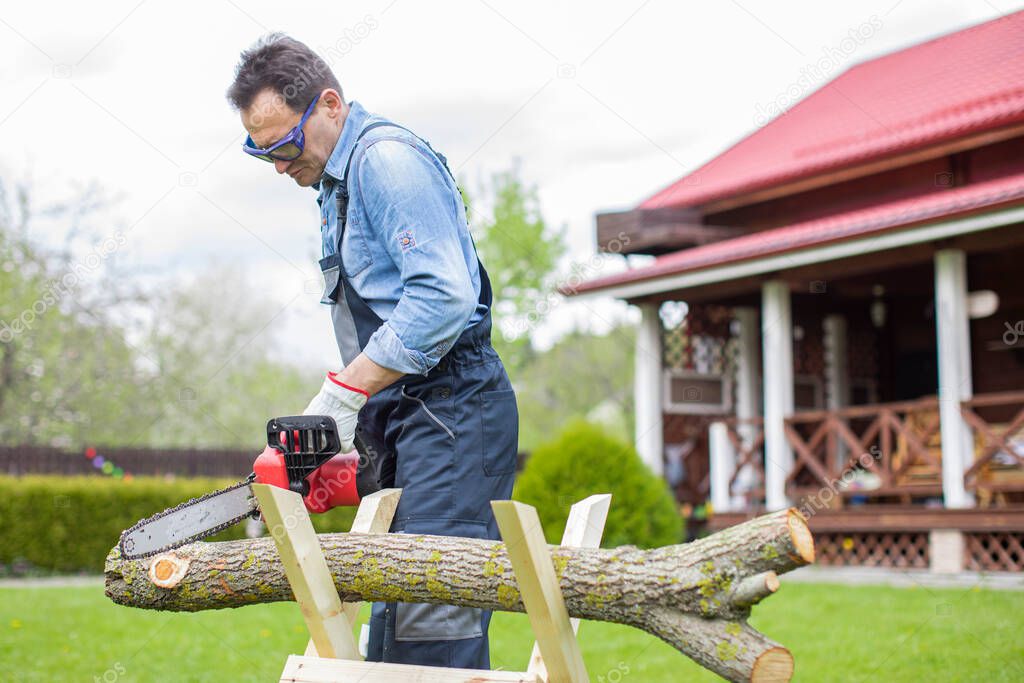 Lumberjack in working uniform sawing tree trunk on sawhorse with electric saw