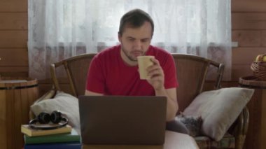 Genç yakışıklı adam kahve içiyor uzaktan bilgisayarla çalışıyor, evde sörf yapıyor.