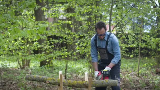 Handsome lumberjack in working uniform sawing wood with chainsaw on sawhorse — Stock Video