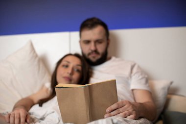 Beyaz pijama giyen iki aşık yatakta iki kişilik bir kitap okuyor.
