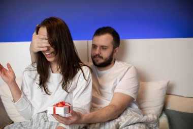 Neşeli adam gülümseyen kız arkadaşının gözlerini avuç içi ve hediye kutusuyla kapatır.