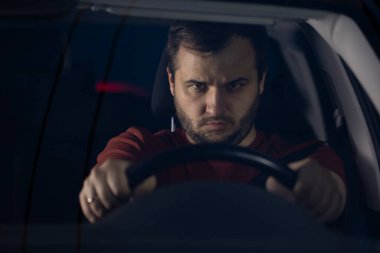 Genç sakallı adam yüksek hızda araba kullanıyor ve direksiyonu elleriyle tutuyor.