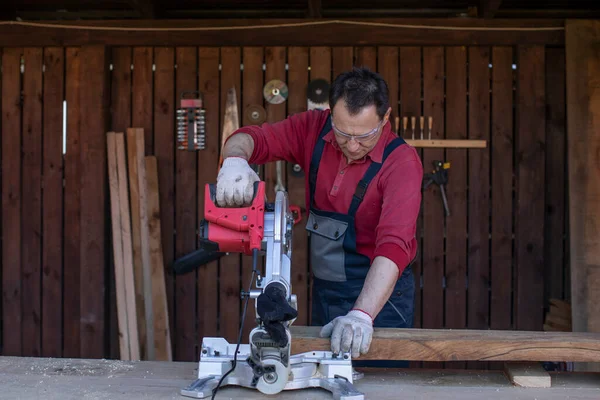 Voksen mand i beskyttende overalls og briller skærer træbjælke ved hjælp af rundsav - Stock-foto