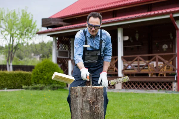 Hombre adulto con camisa vaquera y mono cortando madera con hacha en el campo — Foto de Stock