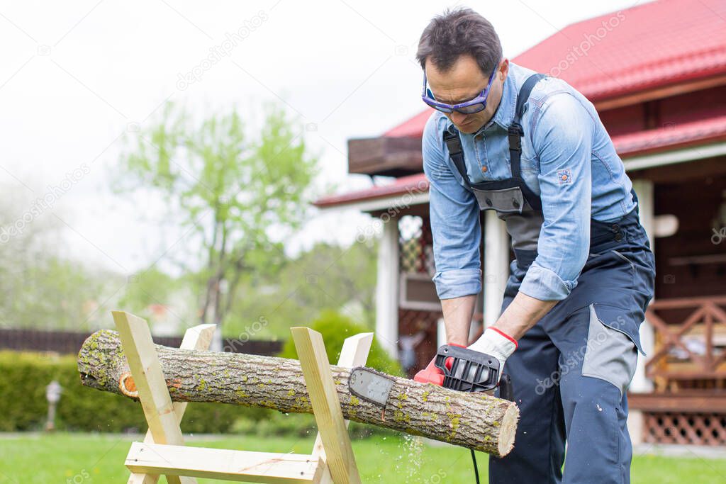 Man in overalls saws a tree on sawhorses in courtyard with a chainsaw.
