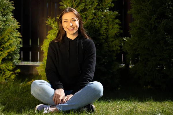 Junge glückliche schöne Frau sitzt auf grünem Gras im Park im Freien blickt in die Kamera — Stockfoto