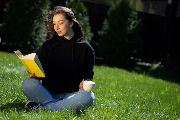 Genç, güzel bir kadın, elinde kitap ve bir fincan çayla çimenlerde oturuyor. — Stok fotoğraf