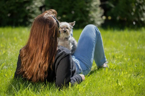 Юная брюнетка в наушниках отдыхает с домашним животным, лежащим на траве в парке — стоковое фото