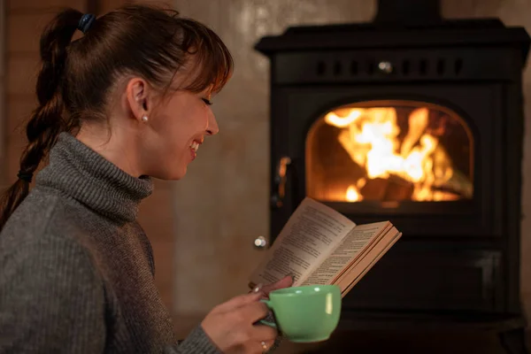 Young woman resting with cup of hot drink and reading book near fireplace