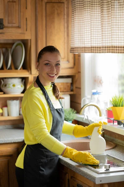 Sorrindo bela dona de casa limpeza cozinha bancada, limpa fogão com spray — Fotografia de Stock