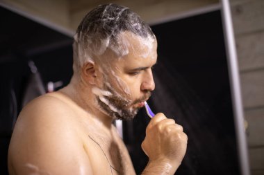 Köpüklü genç yakışıklı adam duş alırken diş fırçasıyla dişlerini fırçalıyor.