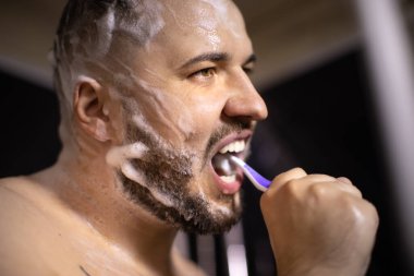 Esmer adam dişlerini fırçalıyor saçını ve sakalını saç bakım ürünüyle sabunluyor.