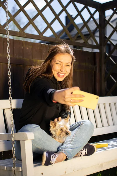 Linda menina sorridente feliz segura um cão em seus braços e toma uma selfie. — Fotografia de Stock